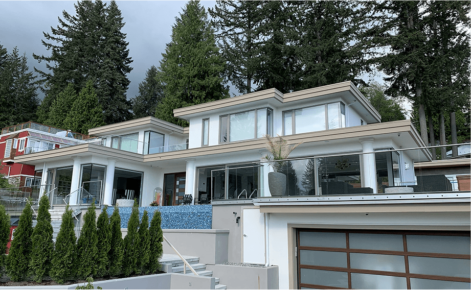 Modern multi-level house with large glass balconies and white siding, surrounded by tall evergreen trees.