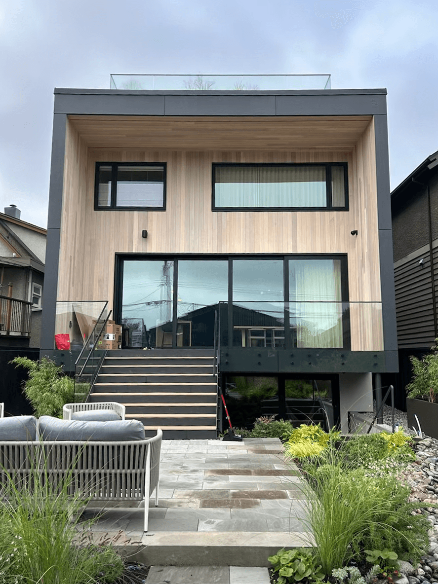 Modern two-story house with wood siding, large glass windows, and a patio garden with outdoor seating.